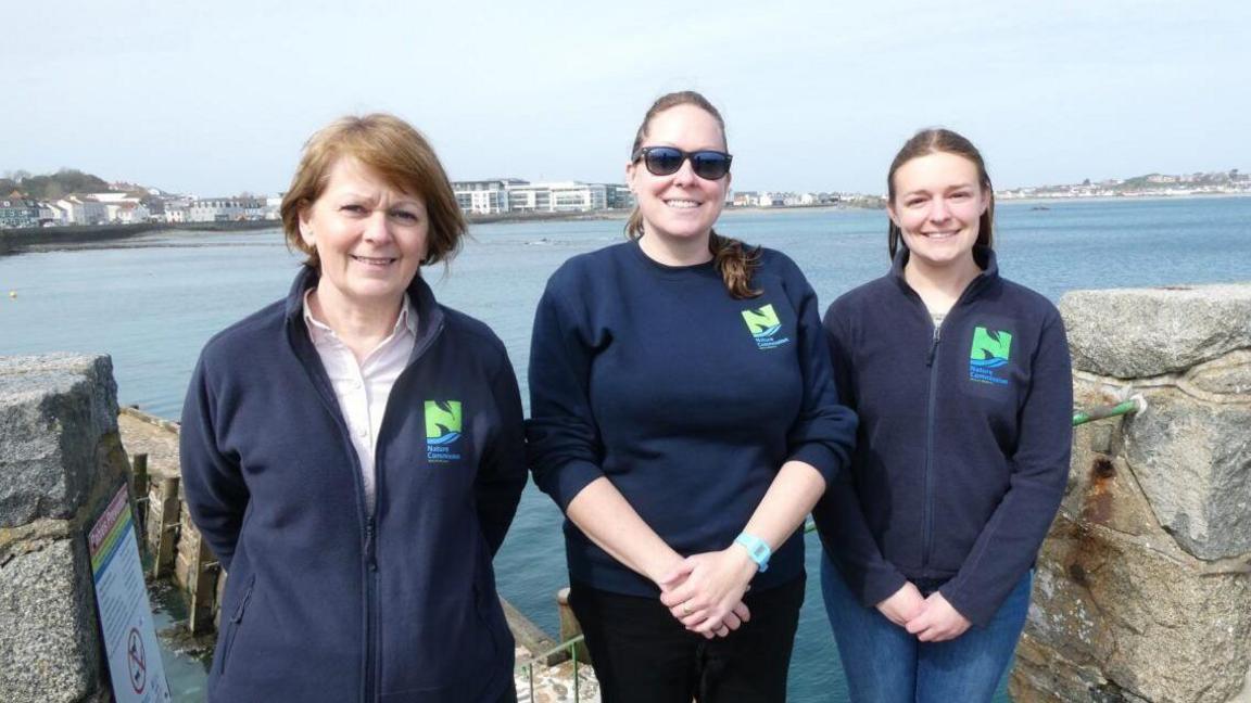 A team photo of Head of Operations and Education Angela Salmon (left), CEO Jessi Jennings (middle) and Ecologist Charlotte Burgoine (right) stood in front of blue sea. There are walls either side of them.