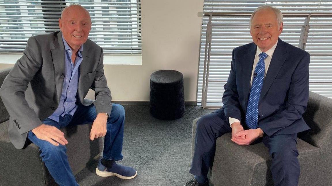 Jasper Carrott with grey jacket and blue shirt is on the left. Nick Owen in a blue jacket and tie and white shirt is on the right. They are sitting down. They are in a room with a grey carpet and white walls.