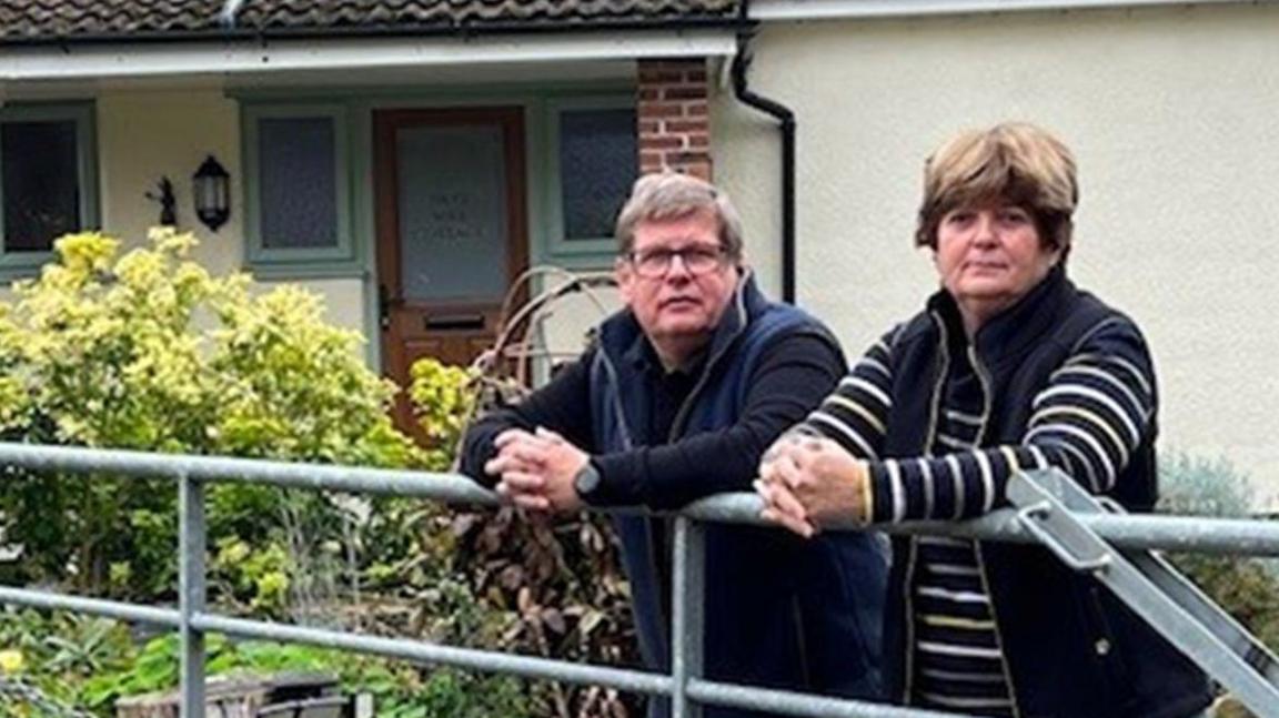 Peter and Catherine Hodgson leaning on the grey gates of Hett Mill Cottage, with the home's brown entrance door in the back.  The house is white and has a small garden with green plants in front of it.  Peter is wearing a black jumper and trousers with blue body warmer and has short grey hair and glasses. Catherine is wearing jeans, a black and white striped jumper and a black body warmer. She has short brown hair. 