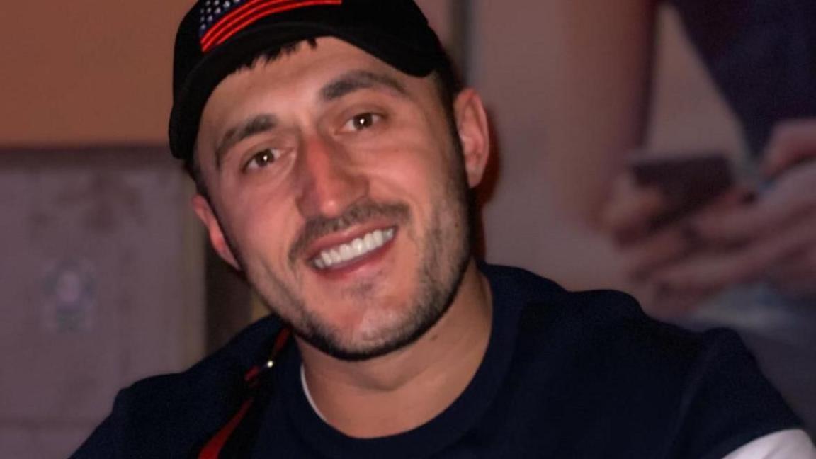 Sali Byberi is smiling at the camera and is wearing a black top and a black cap. He has black stubble.