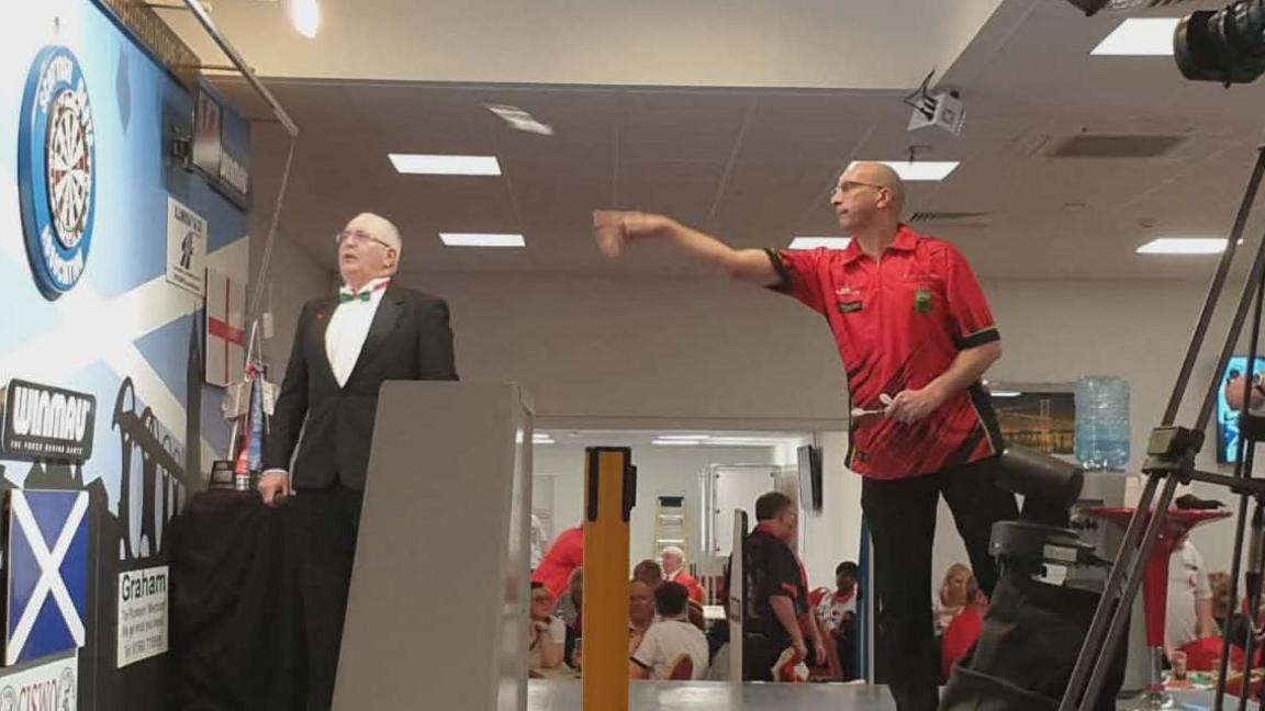 Clive Langford standing at the oche throwing a dart from right to left of the image