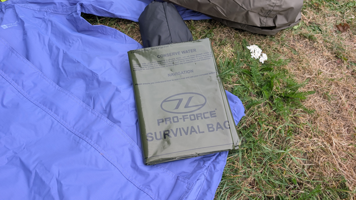 A khaki green survival bag folded on top of a blue waterproof jacket and a bag on the right. The items are laid out on a grassy area.
