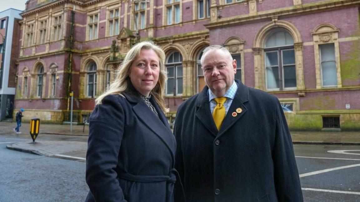 A woman with long blonde hair and a man with short greying hair are both wearing long black coats. They are standing in a city street in front of an ornate brick building with honey-coloured arched windows throughout.