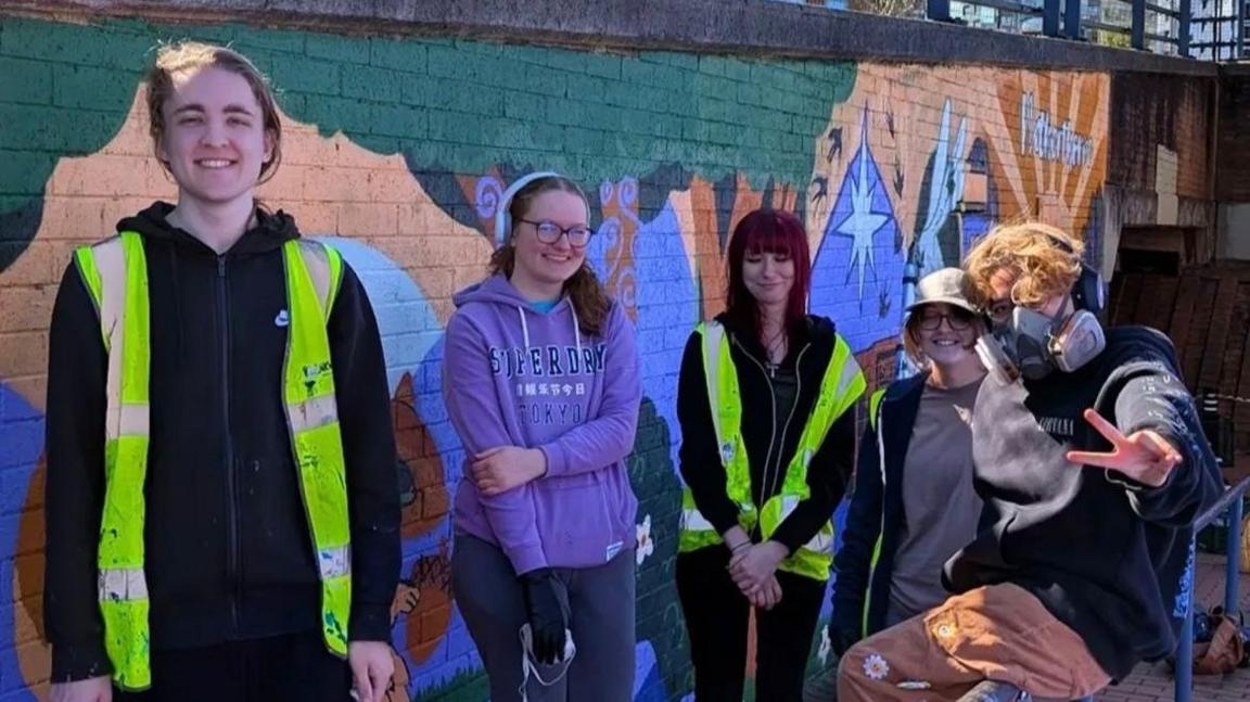 Five people in their late teens or early 20s. They are all smiling at the camera with their work behind them. It looks a sunny day.