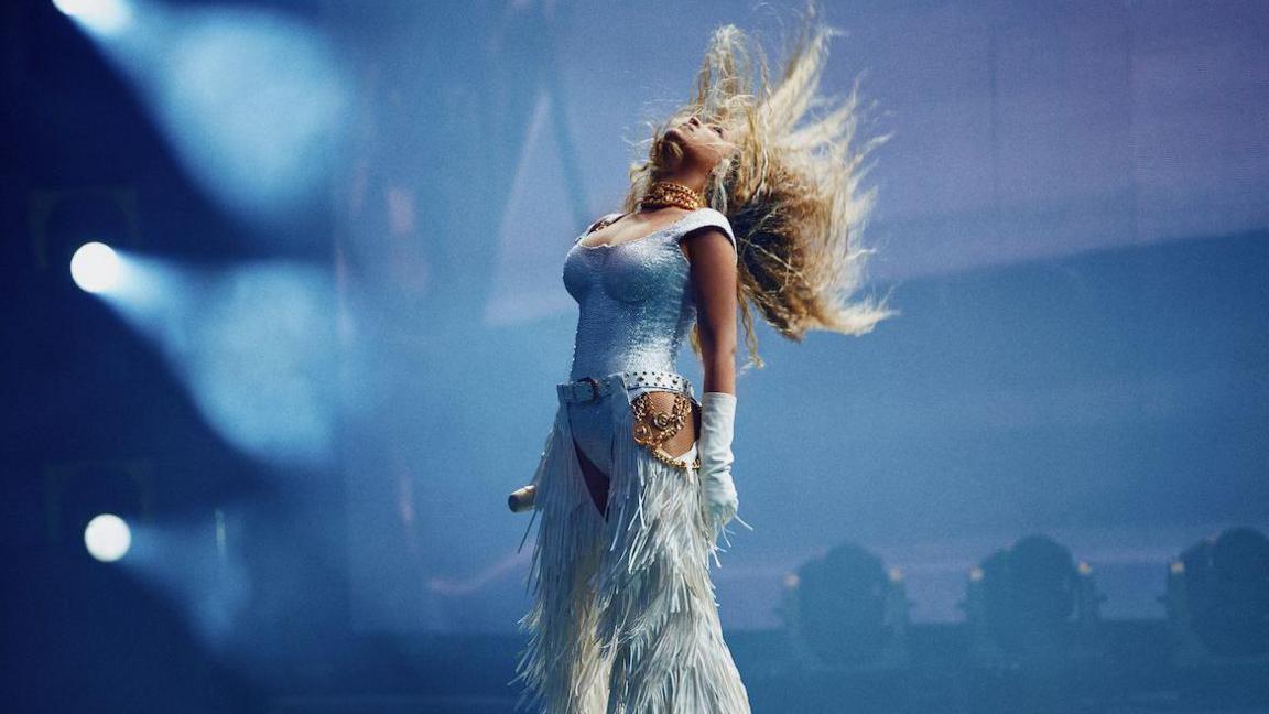 Beyoncé flicks her hair back while wearing an all-white outfit which includes fringed chaps.