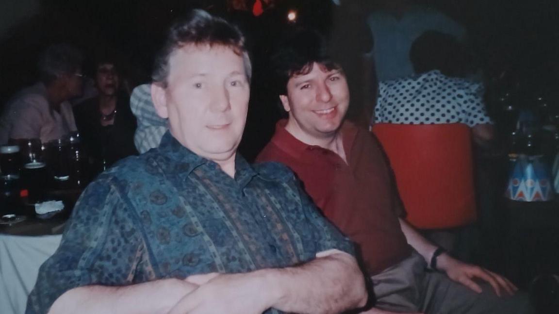 The photo shows two man looking at the camera. The one closest to the camera, on the left, is wearing a blue patterned shirt, and has grey hair. The one furthest away from the camera is wearing a red shirt, and has brown hair. They're sat in a restaurant, and you can see a table with glasses on behind them.