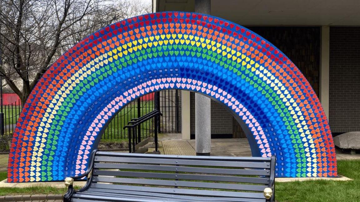 A large memorial in the shape of a rainbow covered in colourful hearts, a bench in the foreground