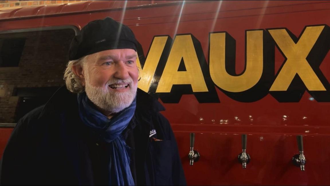 Si King, who has a white beard and is wearing a black cap, standing by beer taps with 'Vaux' on the side.