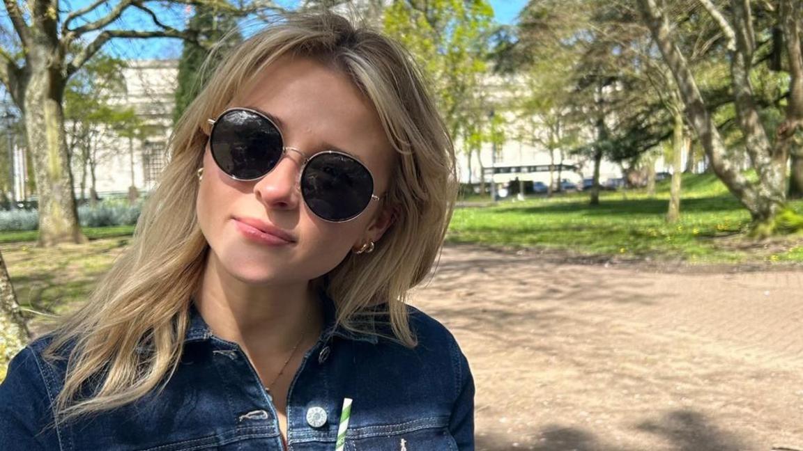 Eve Jones pictured in a dark denim jacket. She wears round sunglasses and looks at the camera. She has blonde mid length hair and wears small gold hoops.