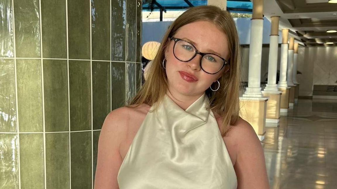 George Smith is smiling and wearing a formal cream-coloured dress. She has long blonde hair and is wearing brown framed glasses. There are a series of columns behind her in the room she is pictured in. 