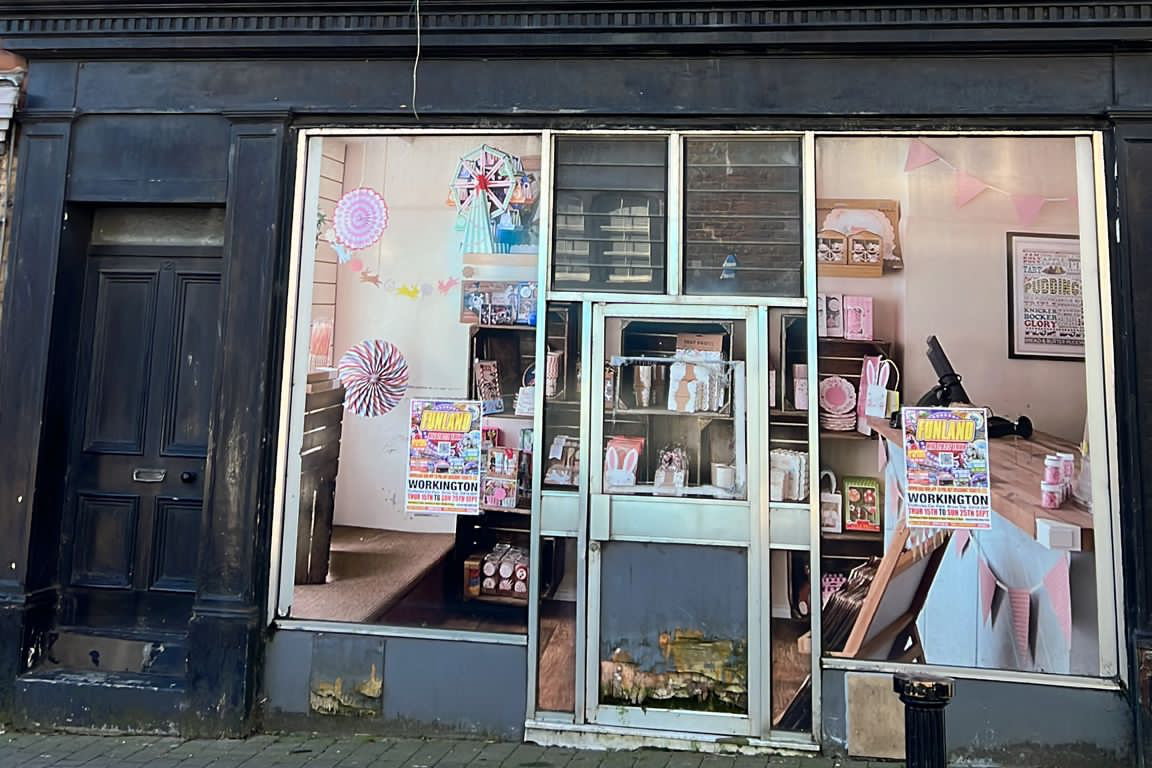 Image of the With Love Photography building before it was refurbished. It has a metal door frame. Along with the window areas on each side, it is boarded up and covered with a large vinyl sticker intended to look like the interior of a shop. Paintwork around those areas is peeling.