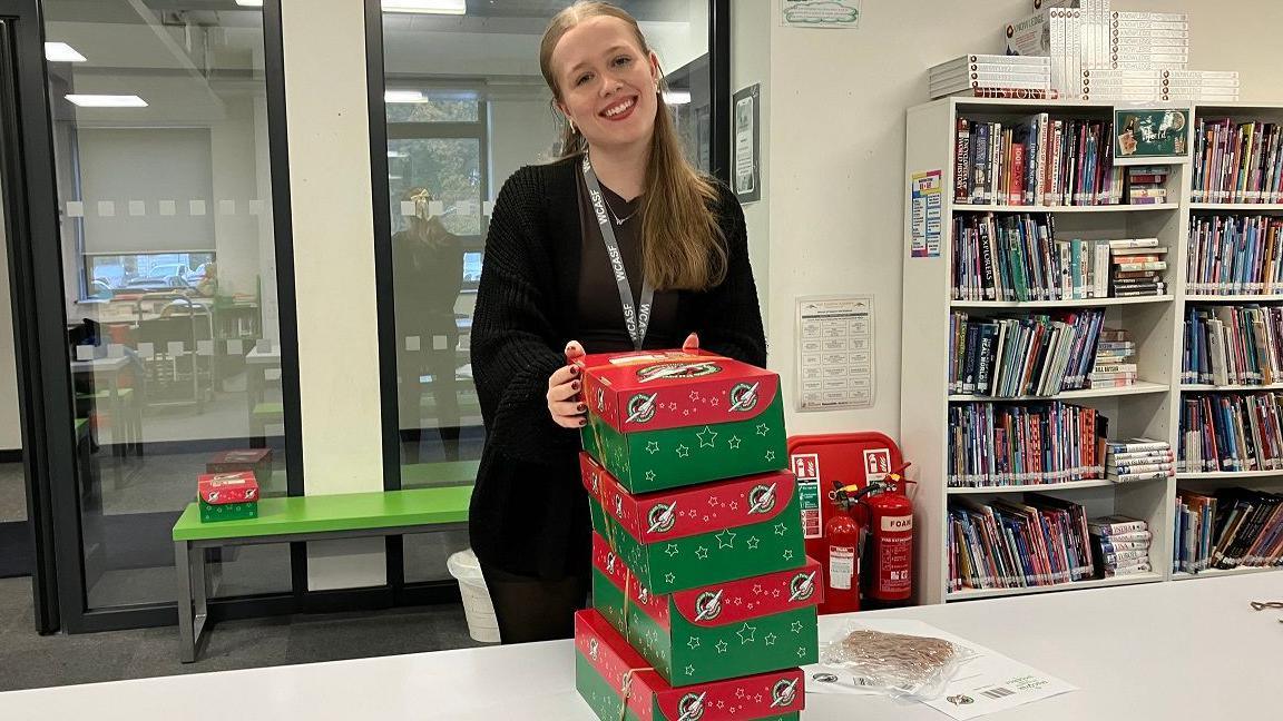 Year 13 student Grace stands in the library with four shoeboxes packed with gifts. She is wearing a black jumper and lanyard and she is smiling. She has long blonde hair.