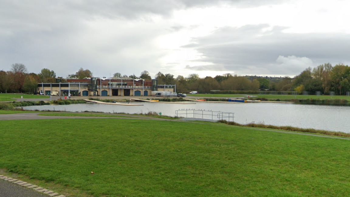 A lake with a large building at the end of it. There is grass surrounding it. On the water there are four yellow floating jetties.