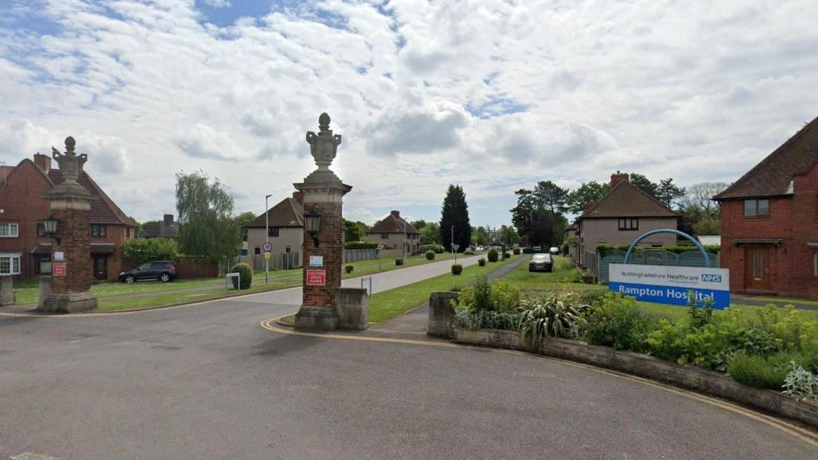 Streetview image of the entrance to Rampton Hospital, showing a large blue NHS sign, then a long road lined with widely sited residential houses.