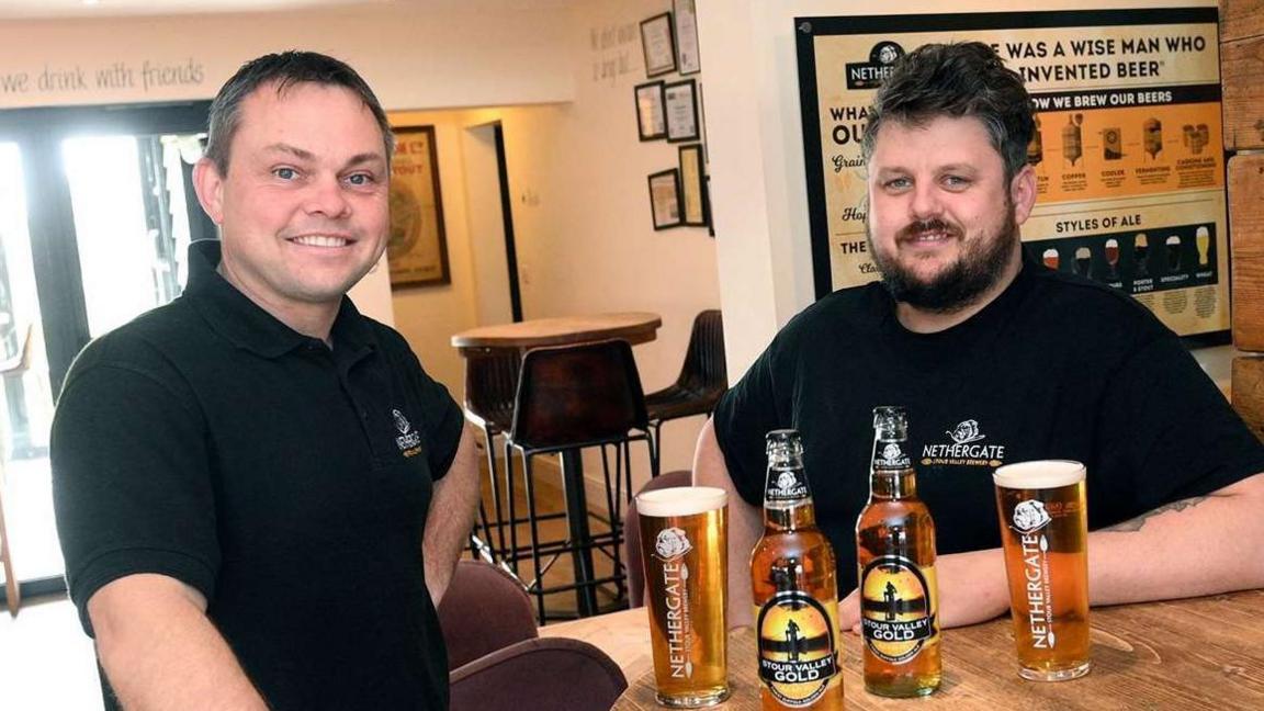 Rob Crawford in a black polo sitting at a table in a pub or bar room, with two pints and bottles at the table, with beer inside. He sits with head brewer Ian Carson who has dark hair and also wears a black polo.