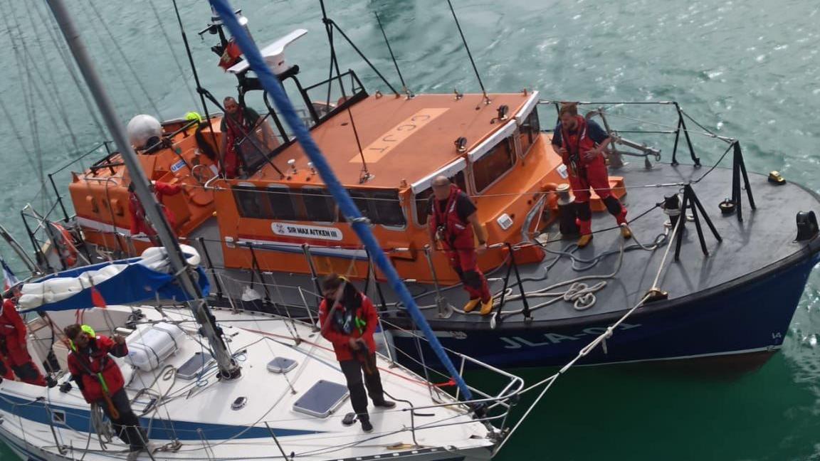 A large orange lifeboat pulled alongside a small white yacht. Crew members stand across both decks. The water is calm.