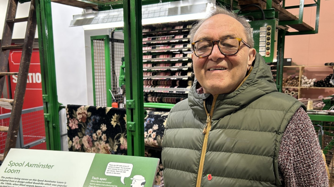 A man with short greying hair is pictured wearing a green gilet and a purple knitted jumper. He is standing in front of racks containing reels of thread and fabric.