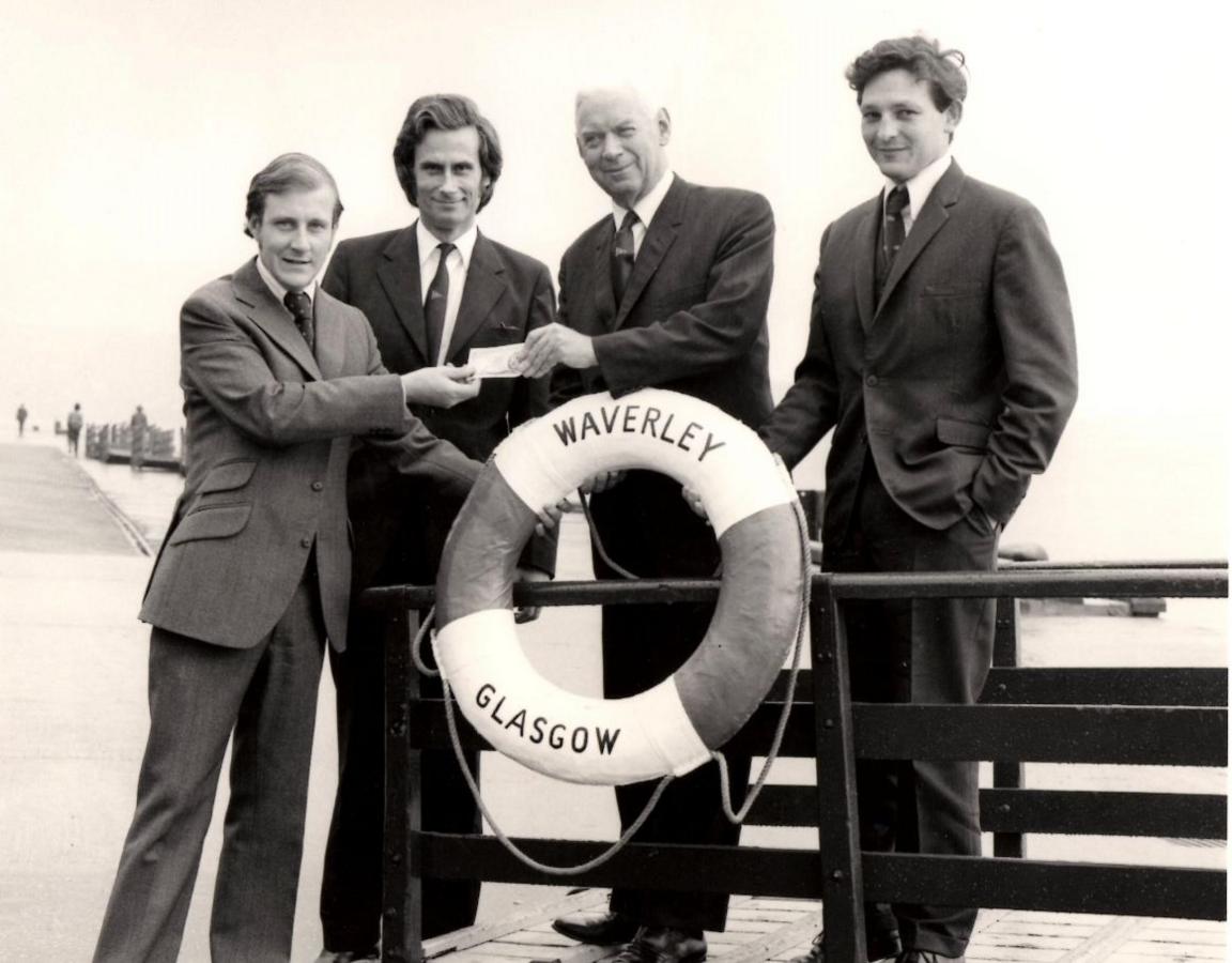 A black and white image of four men in suits, standing around a Waverley life preserver, one is handing over a pound note to an older man.