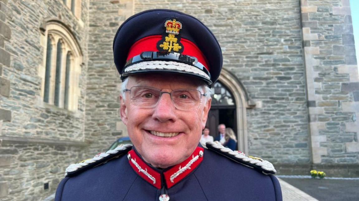 A man, wearing a cap and a navy uniform jacket, smiling at the camera. There is a cathedral building behind him.