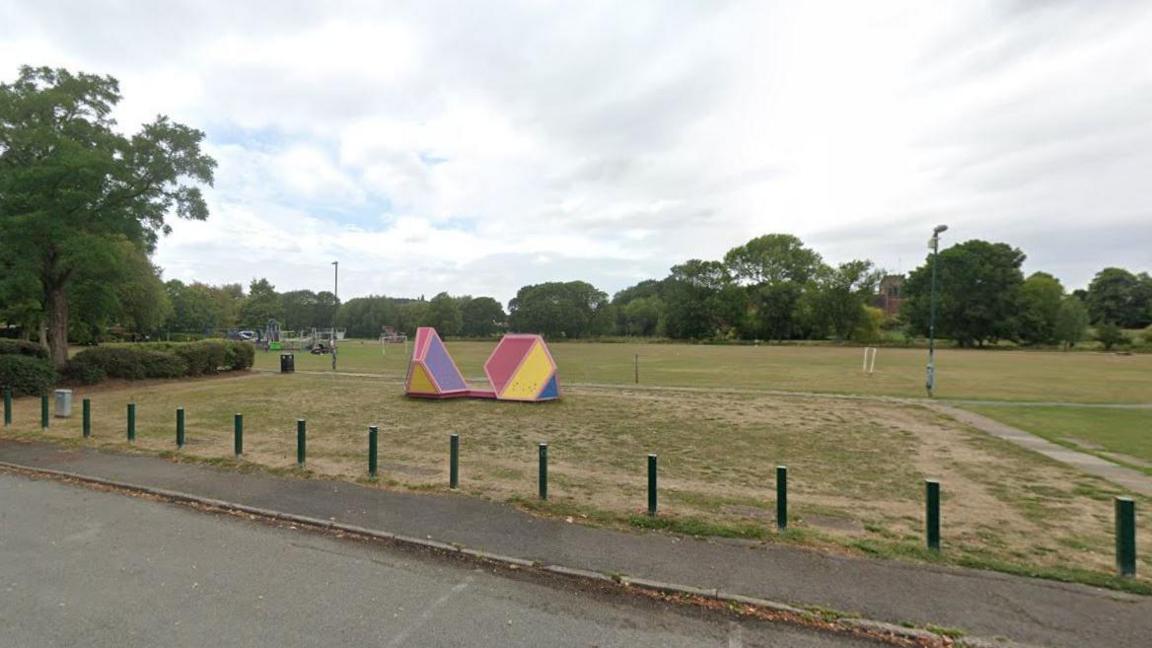 A large green field, in the foreground a pavement and a small area of carpark is visible. There is a red, yellow, purple and blue sculpture/structure in the middle of the otherwise empty field. Trees are visible along the skyline