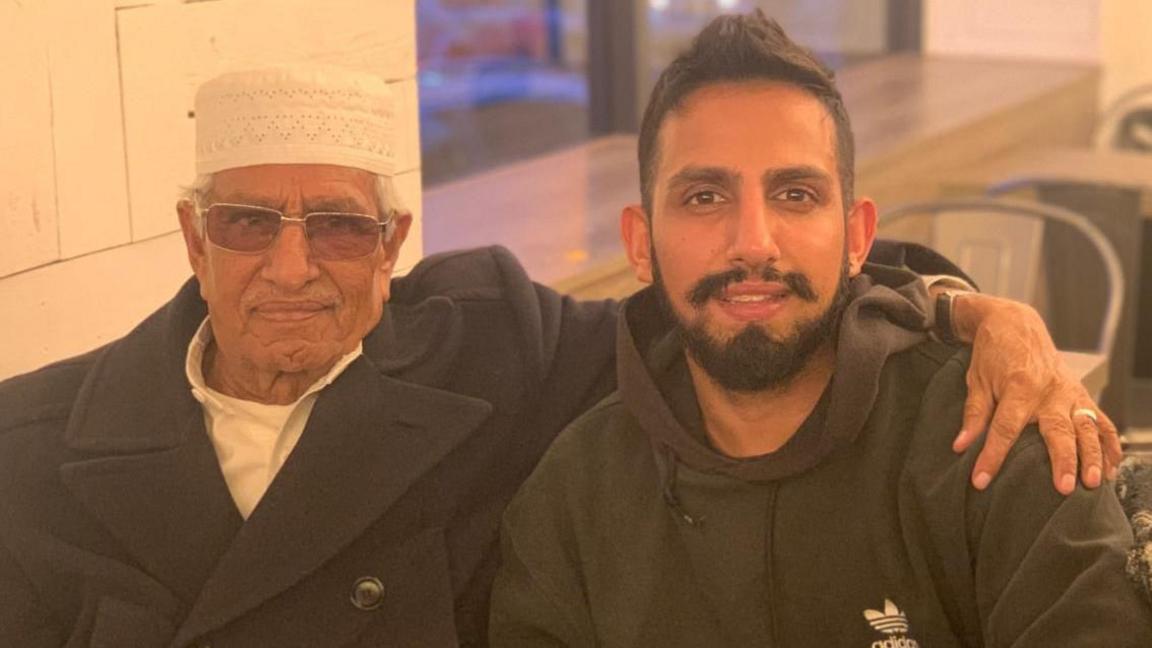 Two men, one older, one younger, sitting together in what looks like a cafe. They smile gently into the camera. The older man has his arm around the younger man's shoulder in a loving way. The older man wears a skull-cap type hat, and the younger man wears a black hoodie.