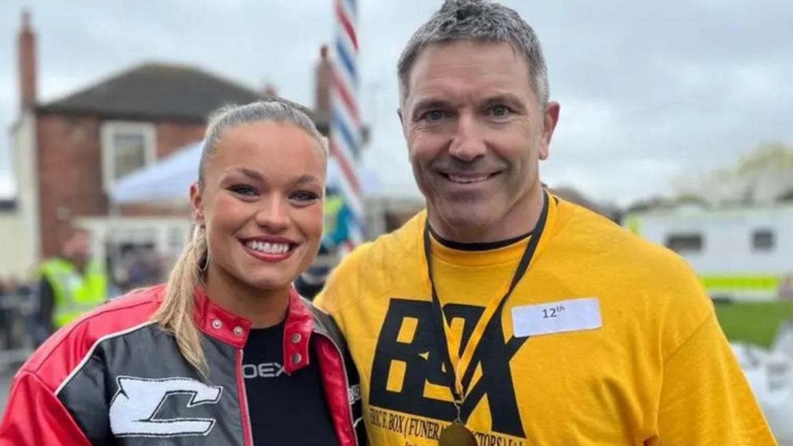 A young woman in a red jacket stands next to a man who is wearing a yellow t-shirt and a medal around his neck.