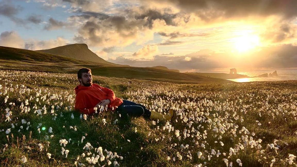 Man with beard and wearing red hoodie, dark trousers and yellow wellies, lying amid flowers in a field with hands clasped, and island coastline in background and sea, with sun glowing brightly in the sky.