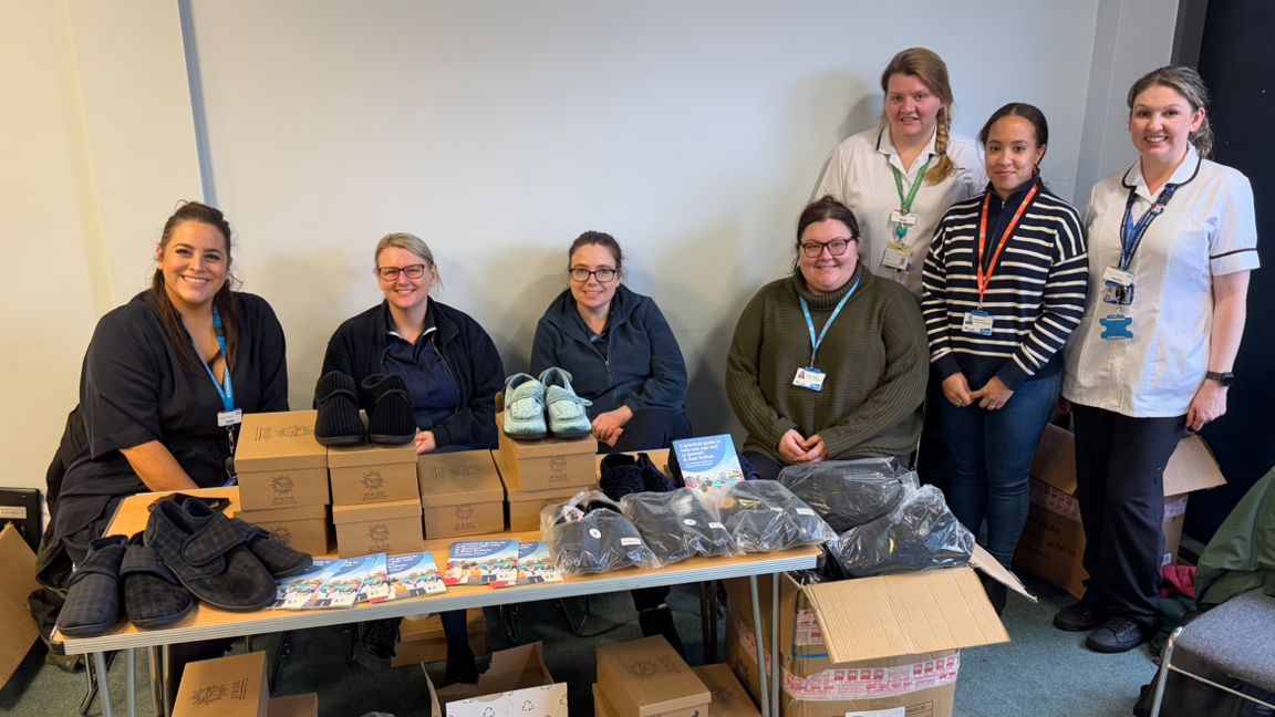 Seven women standing behind a table on which there are multiple boxes of slippers for elderly people.