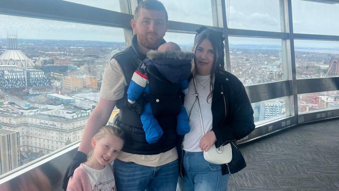 Morgan and Ellie smile for the camera on a viewing platform overlooking Liverpool. Morgan has a baby strapped to his chest and their daughter is standing beside him in a pink cardigan