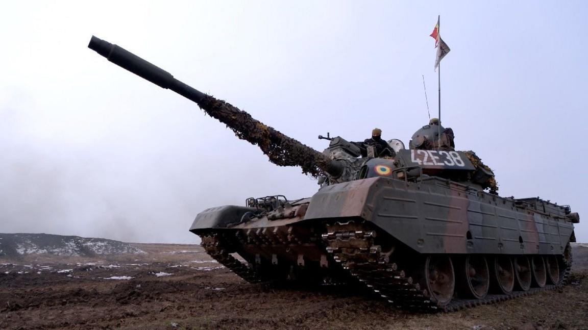 A brown and green military tank moving across a dirt path with a flag on top and its gun barrel pointed upwards