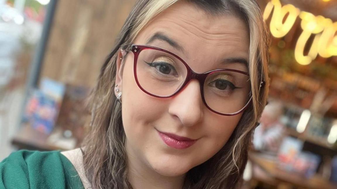 Selfie of Georgia Louise Edwards who has glasses and wavy brown hair is wearing a green top