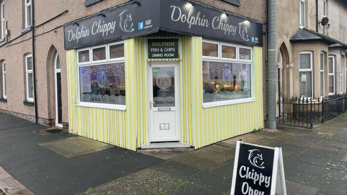 External view of Dolphin Chippy which has a black sign and yellow walls and white windows and door and sits on the corner of Blakiston Street. There is a black and white A-board sign outside saying Chippy Open.