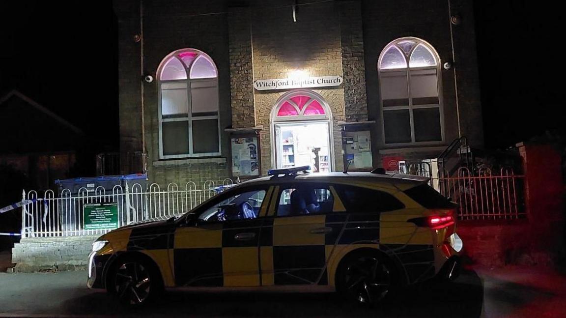 In front of the police cordon on a dark evening is police car. Behind it the door into the post office remains open with the lights shining bright inside. Above the doorway is a sign which reads Witchford Baptist Church.