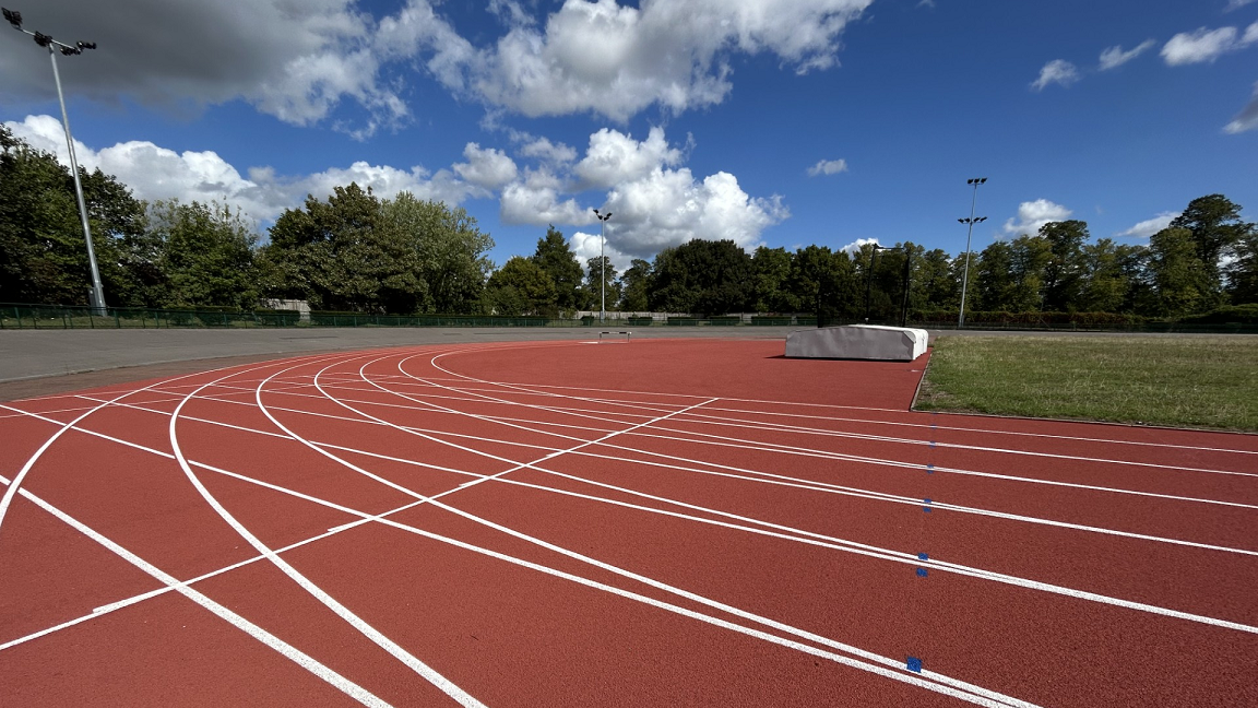 The corner of the orange athletics track under a blue sky. There's floodlights at the edge and trees behind it.