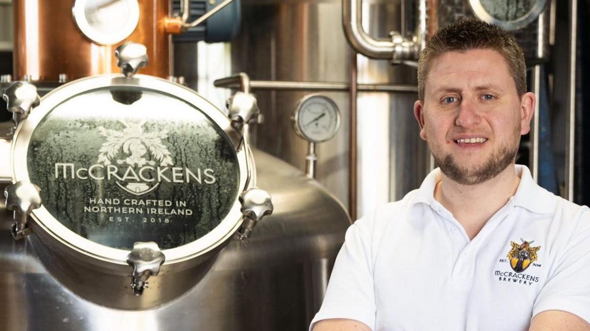 Ryan McCracken standing beside his beer fermentation tanks, with glass reading 'McCrackens Hand Crafted in Northern Ireland Est. 2018'. Ryan has short brown hair and a beard, he is wearing a McCrackens embellished white polo shirt and smiling at the camera. 