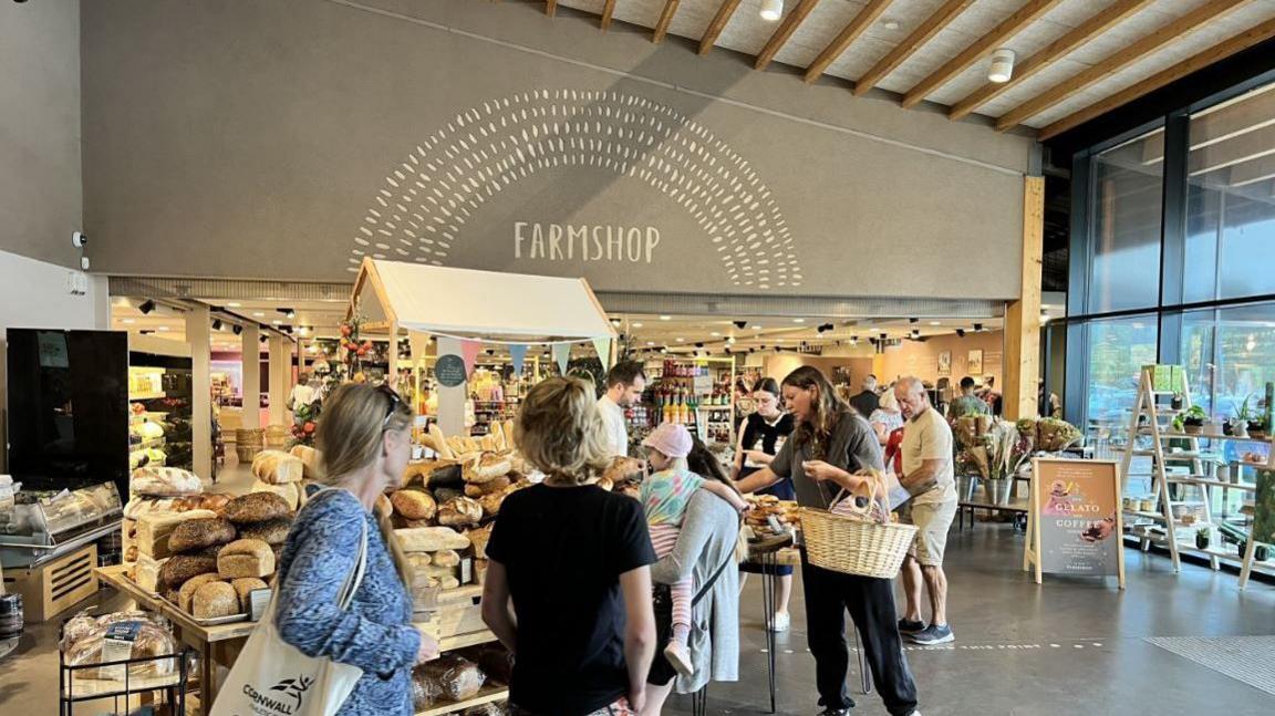 People inside Gloucester Services look at products on display. They are carrying baskets and shopping bags, and a large fresh bread display is visible in the foreground