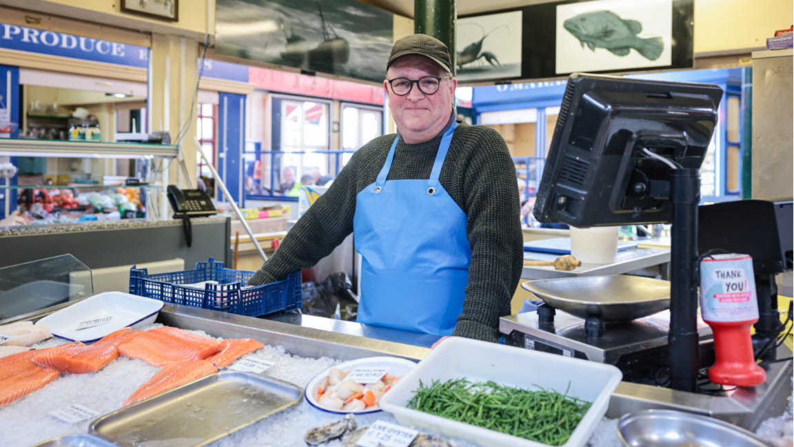 Dave Walker, wearing a blue apron and green jumper, smiles behind the counter with salmon and other fish in front of him