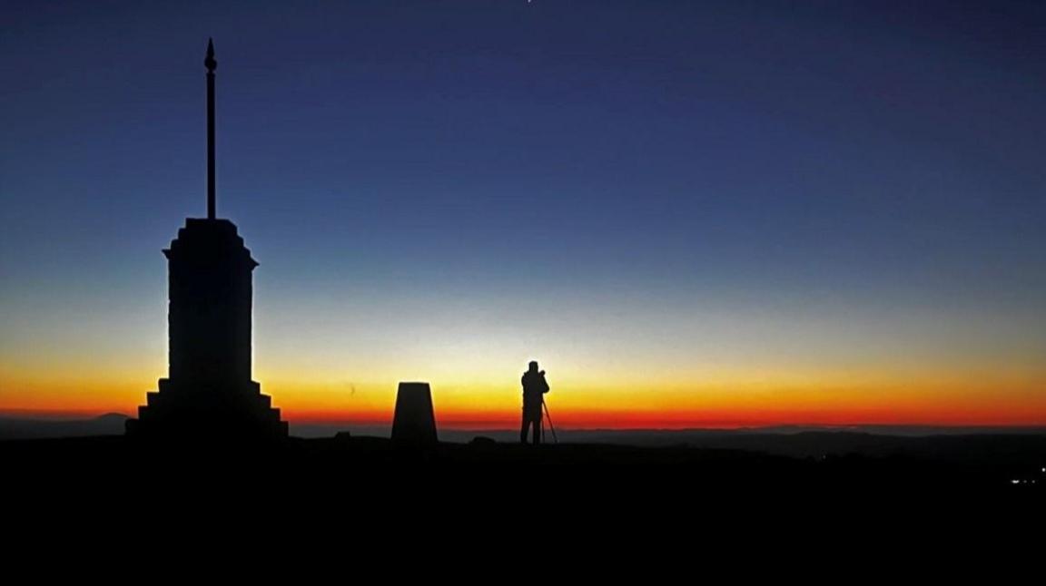 A person silhouetted on a dark night, with a telescope. A reddish/orange tint is on the horizon.