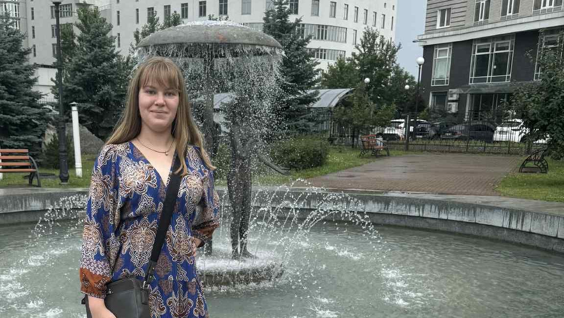 Sasha Shevchenko stands by a fountain which has a mushroom with figures underneath. She is wearing a blue dress and has a handbag over her shoulder. She has long blonde hair with a fringe and is wearing a necklace.
