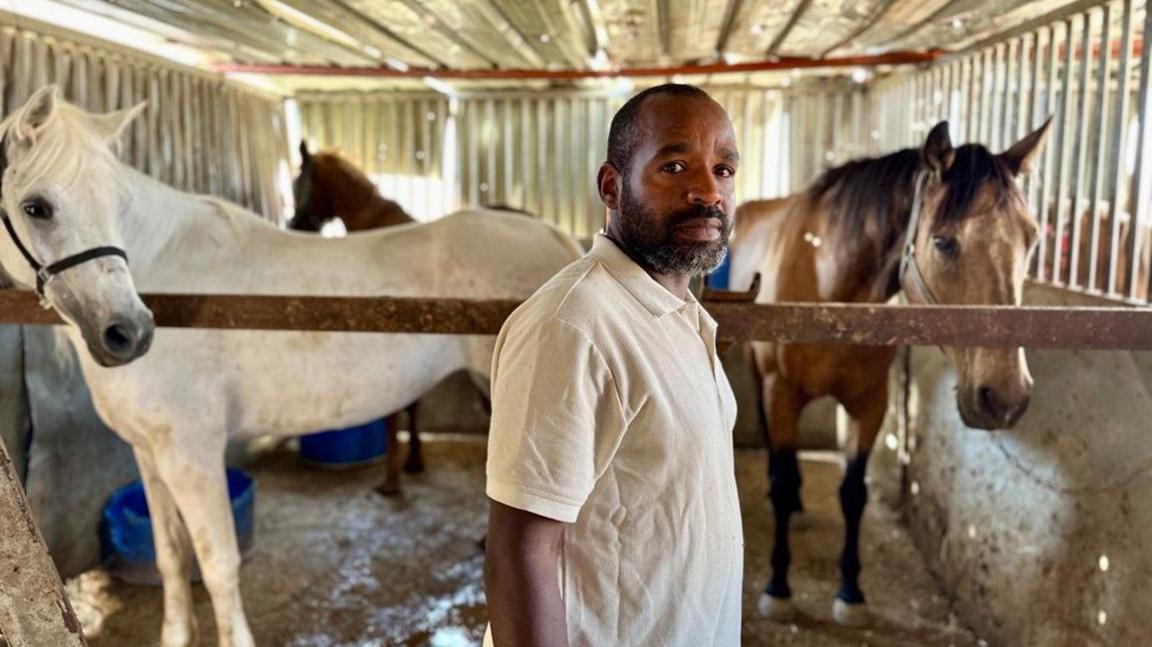 Khaled stands in front of three horses inside a stable