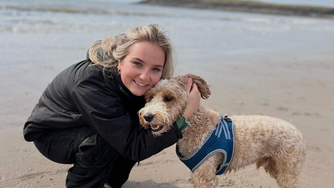 Emily is on a breach hugging a terrier type dog. She has long blonde hair swept back and is wearing a black windbreaker jacket and matching trousers. She is smiling and holding her cheek against the top of the dog's head. 