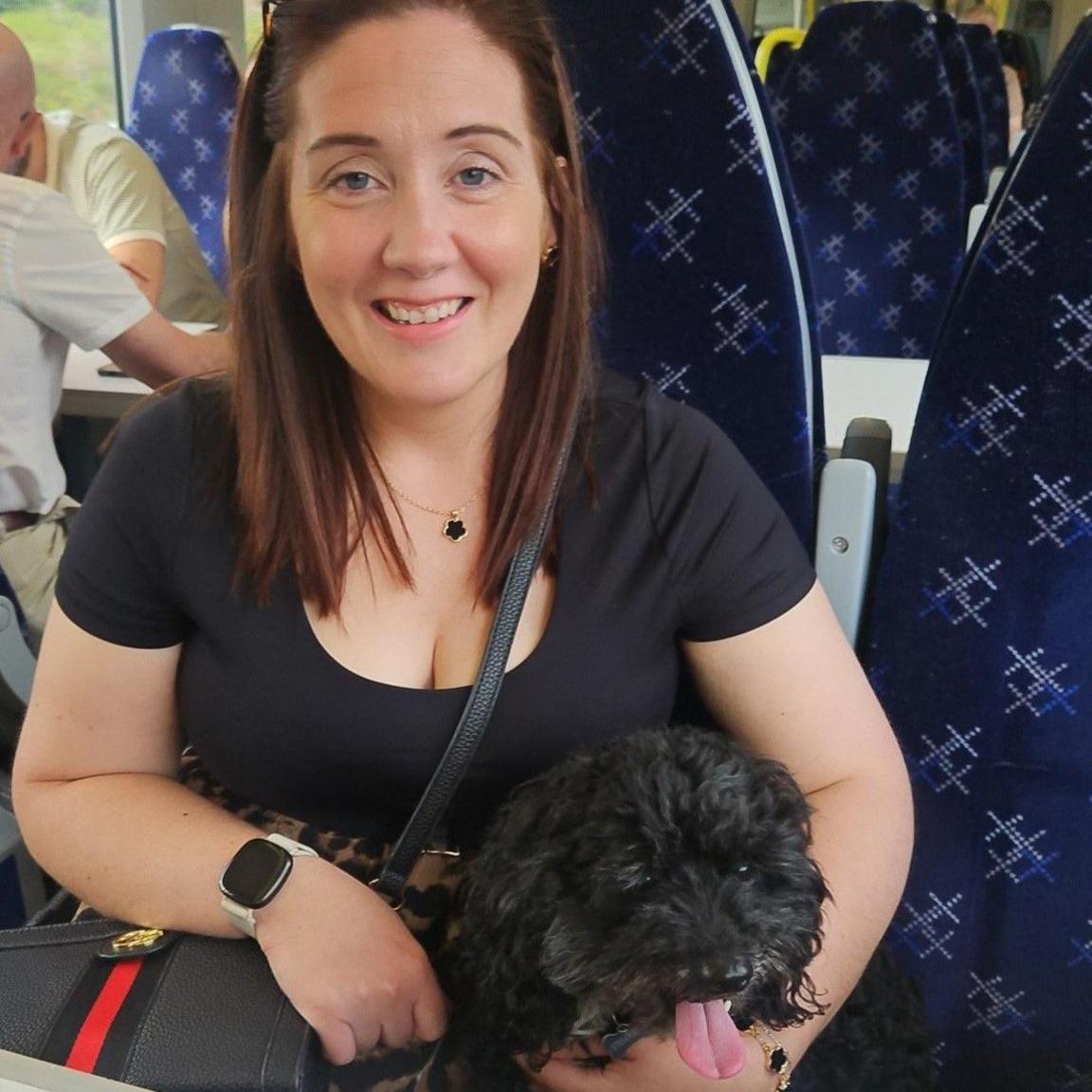 A woman with brown hair smiles while holding a black dog with its tongue sticking out.
