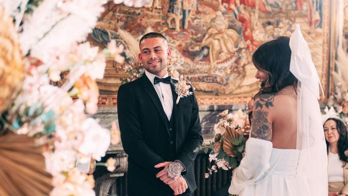 Sionainn Carmichael and Ryan Livesey are photographed on their wedding day. Ryan is wearing a black suit with a peach button hole, and Sionainn is wearing a white wedding dress with a veil. They are both stood facing each other and smiling. 