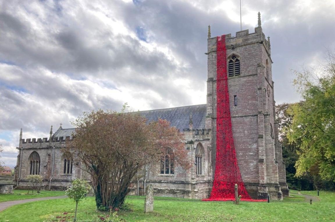 A cascade of poppies on the church bell tower going from the top to the ground. The rest of the church is behind the tower and there are trees in the foreground and to the right.