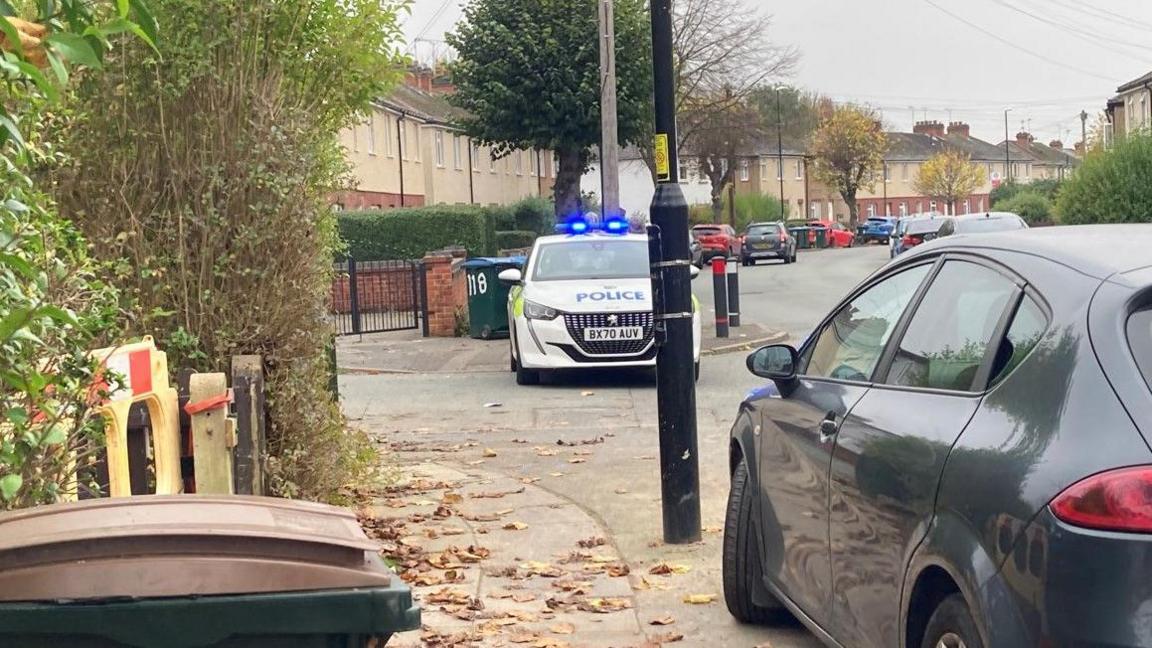 Cars are parked on both sides of the road. A police car is on the left with blue lights on and a bin is in the foreground.