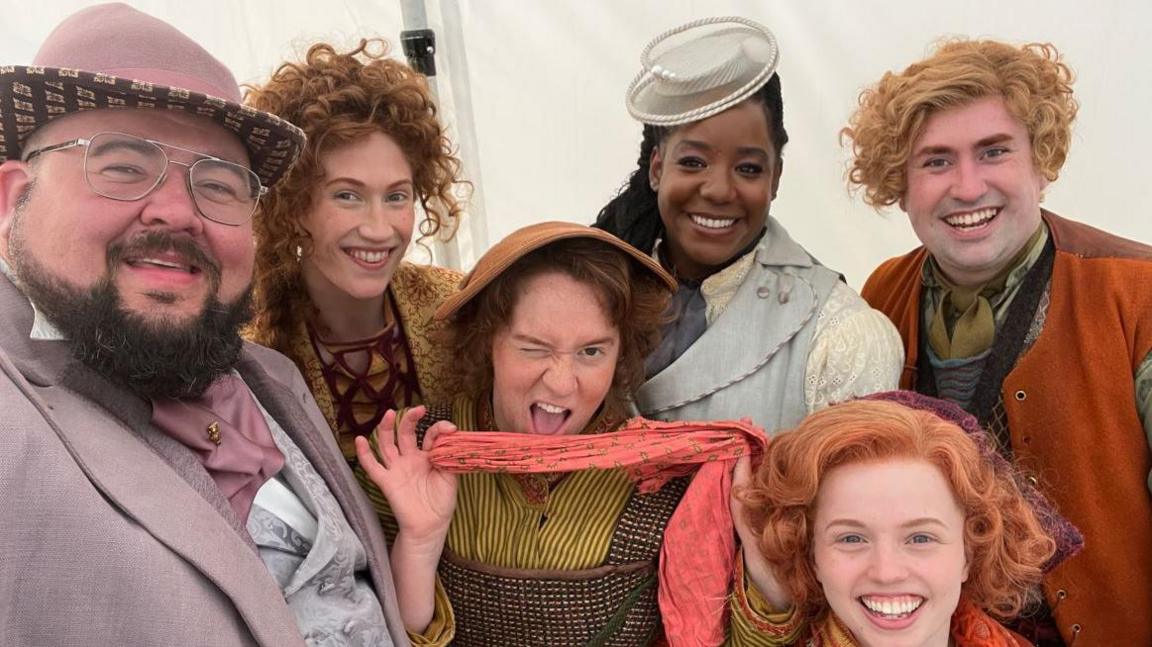 A gathering of people in brightly coloured Victorian-style costumes with wavy hair and hats