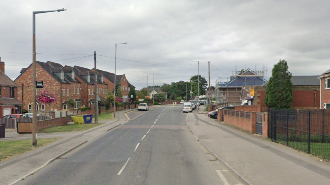 Shafton High Street, with houses and streetlights on either side of the road. On the right-hand side is a Premier shop. Past that, cars are parked half on the pavement and half on the road.