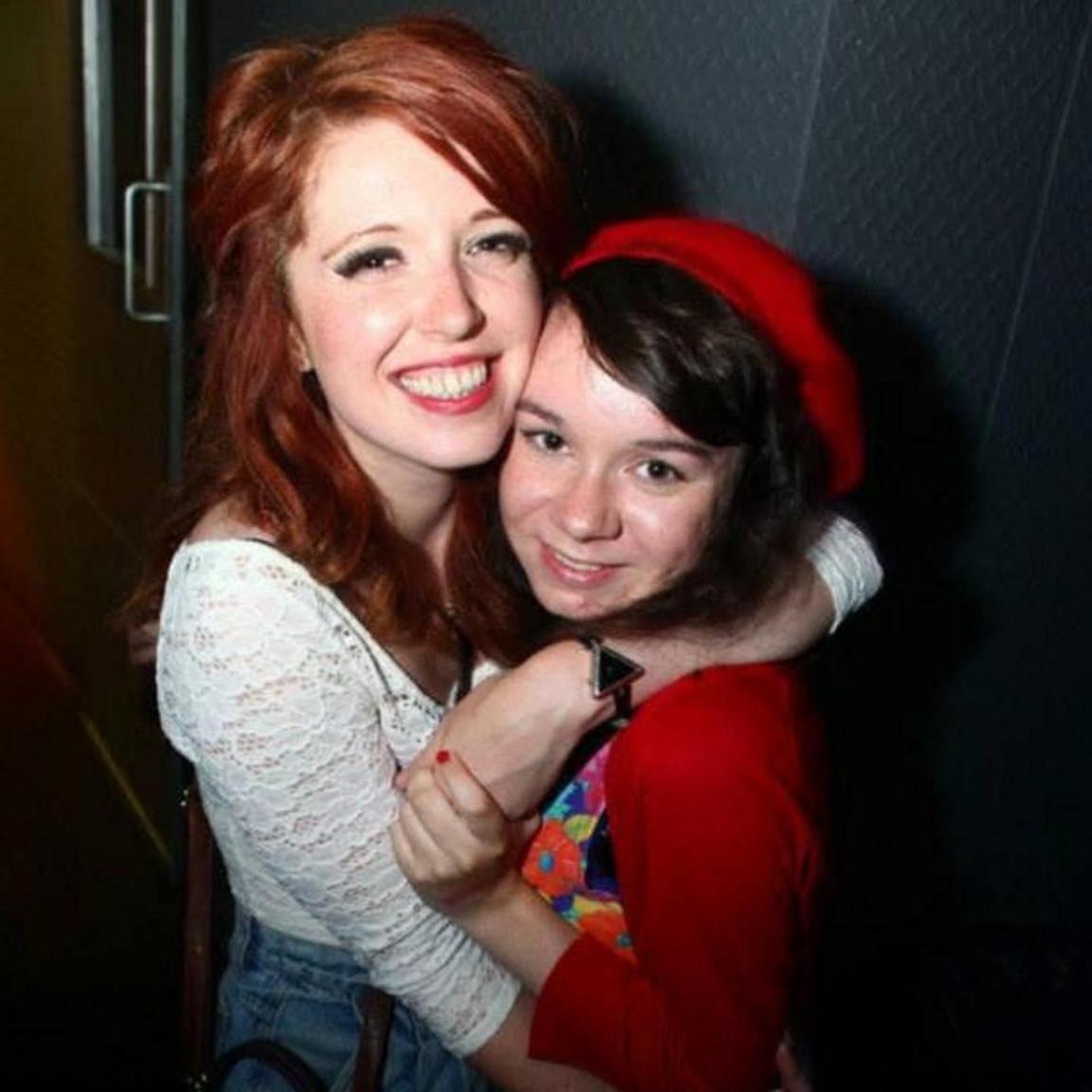 A young woman with red hair has her arm around her friend, who has dark brown hair and wears a red beret. They both smile at the camera.