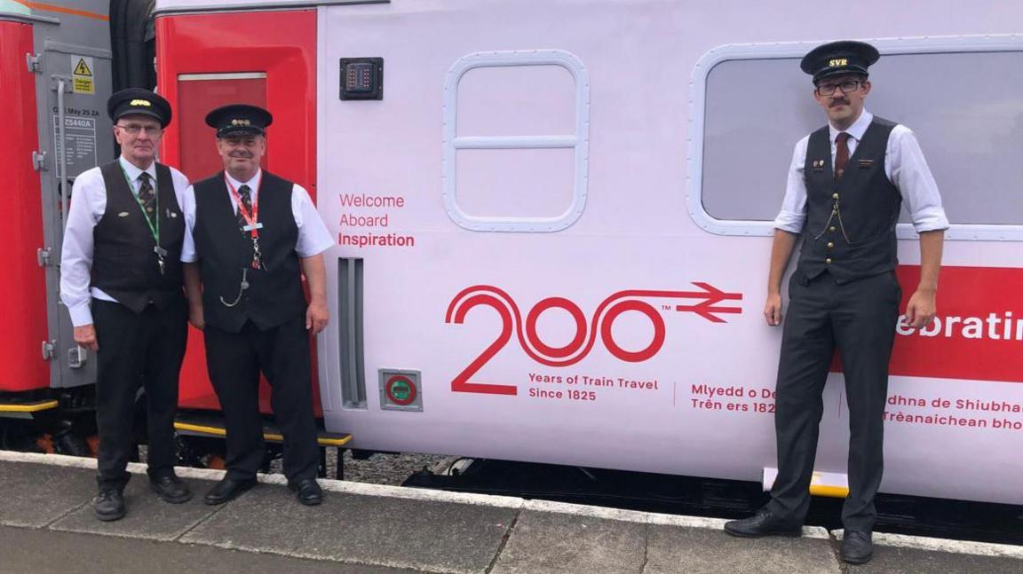Three men in black and white uniforms with black caps standing beside a white railway carriage