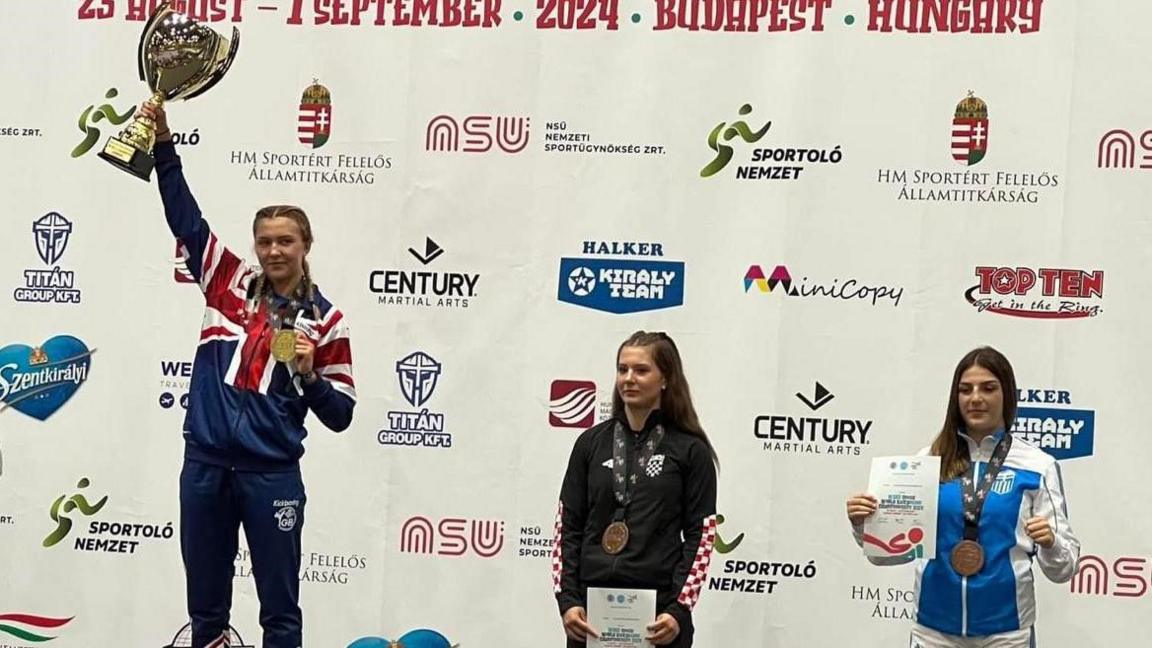 Freya Holmes is stood on the winning podium. She is wearing a red, white and blue GB tracksuit and is holding a large gold cup aloft in one hand. In the other hand, she is holding a large gold medal. Two women are stood next to her in tracksuits, both with medals and certificates